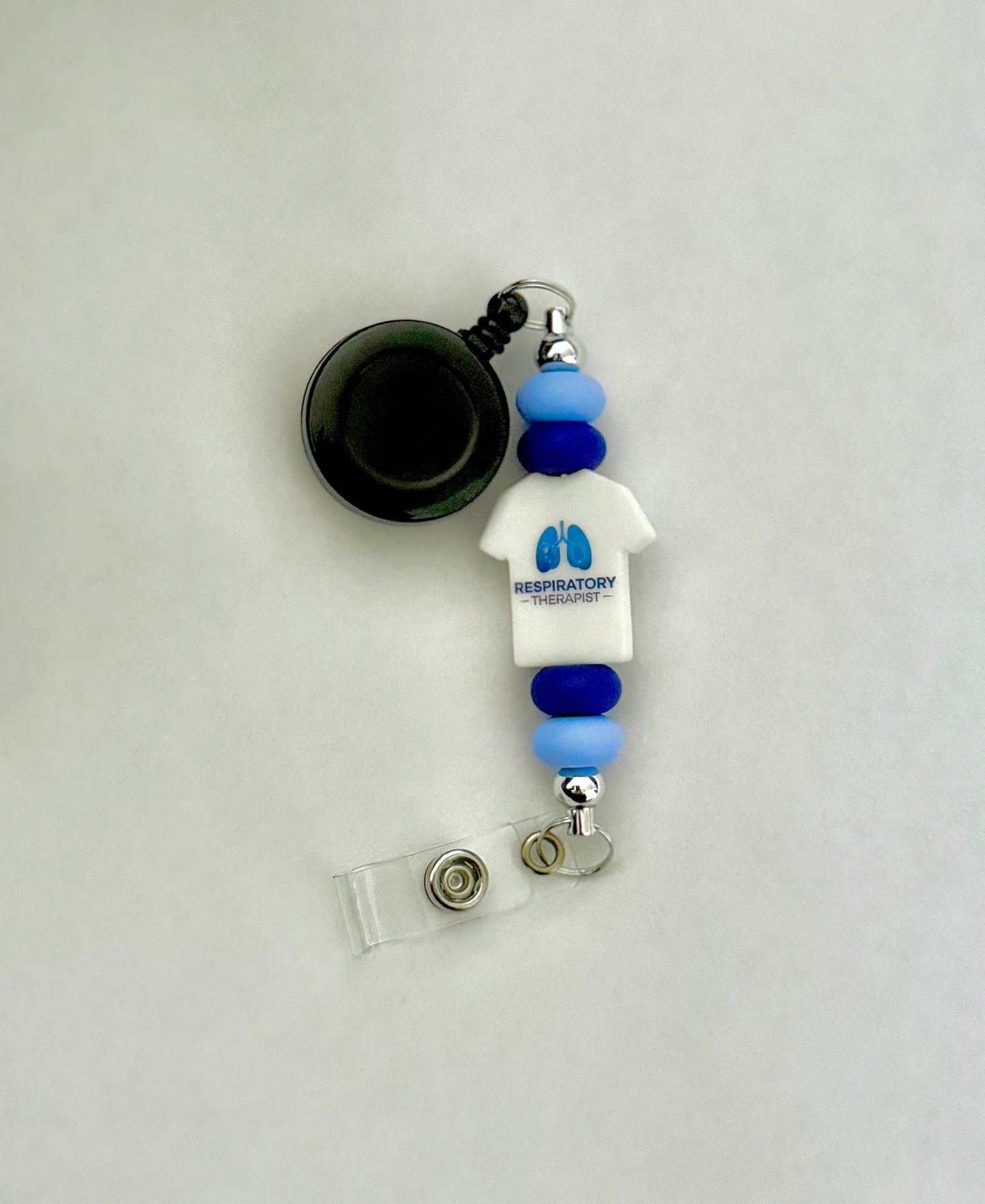 Handmade badge reel with a t-shirt shape focal bead that is white with blue print, light and dark blue silicone beads. Perfect for respiratory therapists working in the medical field 