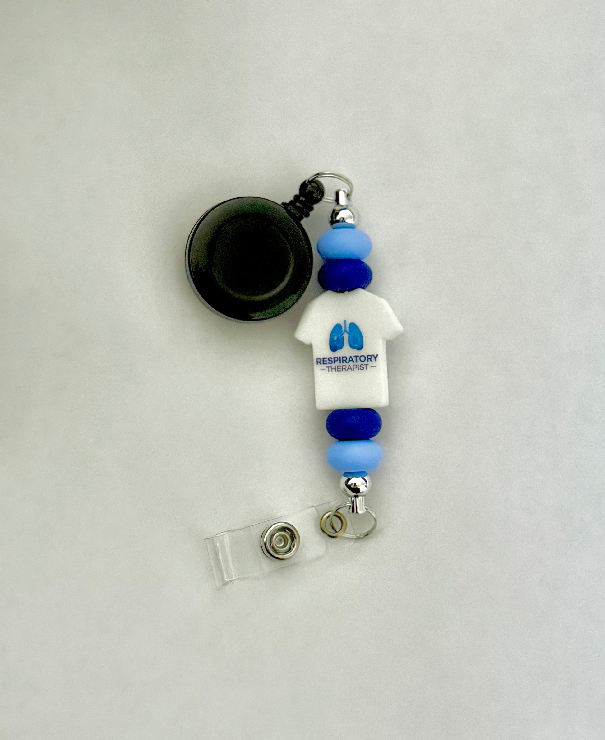 Handmade badge reel with a t-shirt shape focal bead that is white with blue print, light and dark blue silicone beads. Perfect for respiratory therapists working in the medical field 